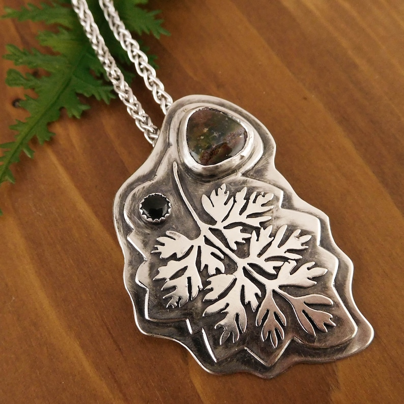 Pu&ograve; includere: Una collana con un ciondolo in argento che presenta un designo dettagliato di foglia di felce con una pietra verde e marrone e una pietra nera.