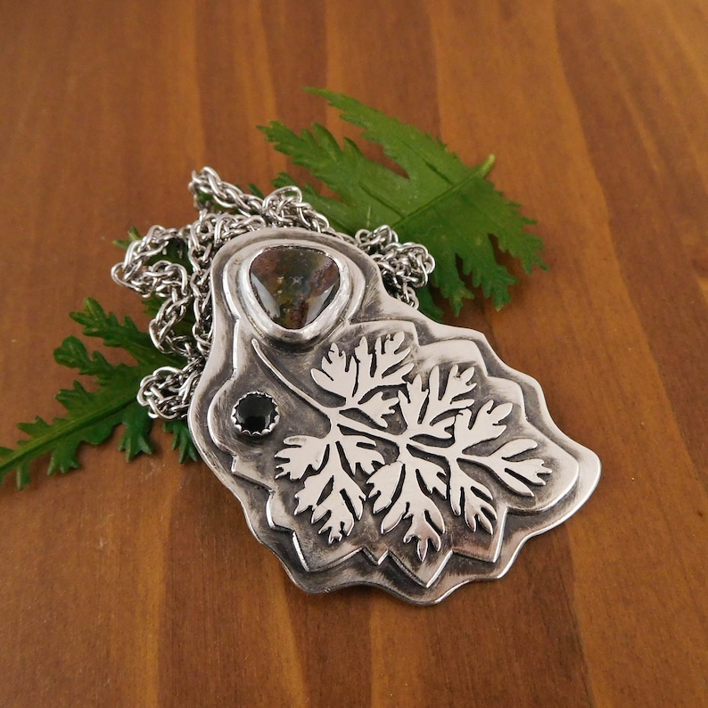 Pu&ograve; includere: Una collana con un ciondolo in argento che presenta un designo dettagliato di foglia di felce con una pietra preziosa verde e marrone e una pietra preziosa nera.