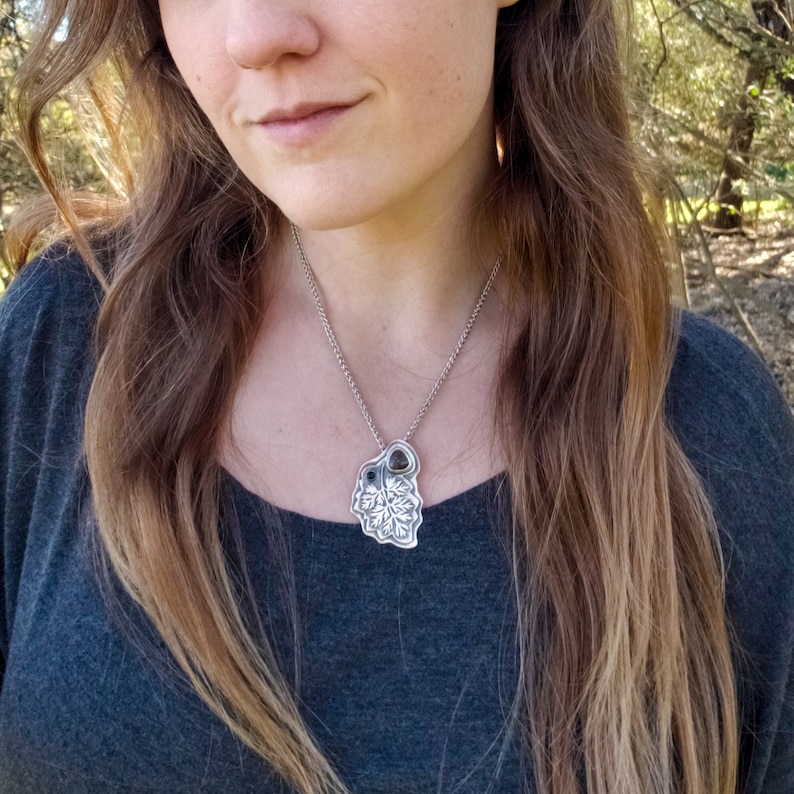 Pu&ograve; includere: Una collana d'argento con un ciondolo a forma di foglia dettagliato con due pietre verde scuro. La collana &egrave; indossata da una persona con i capelli lunghi e castani.