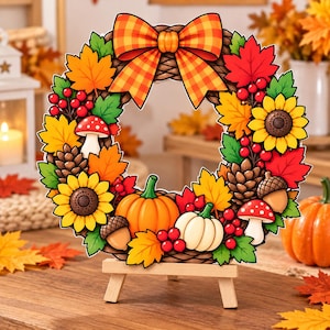 May include: A circular autumn wreath featuring a large orange and yellow plaid bow at the top, decorated with sunflowers, pumpkins, pinecones, red berries, mushrooms, acorns, and colorful maple leaves, displayed on a small wooden easel stand.