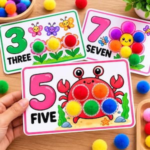 以下が含まれることがあります： 数字とそれに対応するカラフルなポンポンが描かれた3枚の長方形の知育カード。カードには、花が描かれた数字の3、花が描かれた7、カニが描かれた5があり、それぞれ数字の単語が書かれています。