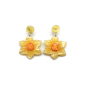 Puede incluir: Un par de pendientes colgantes de flores amarillas con una flor naranja más pequeña en el centro. Los pendientes están hechos de resina y tienen un acabado brillante.