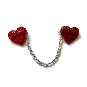 May include: Two red glitter heart charms linked by a silver chain. The hearts have a textured, sparkling finish. The chain is made of small, interlocking links. The background is plain white.