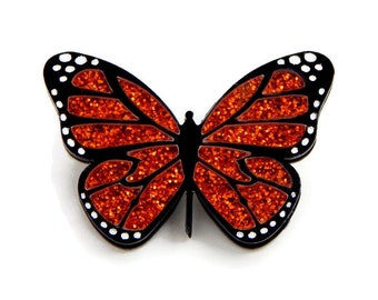 May include: A black and orange glitter butterfly pin. The butterfly has black outlines and white dots around the edges of its wings.