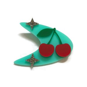 May include: A turquoise blue boomerang shaped brooch with two red cherries and two silver glitter stars.