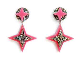 May include: A pair of pink star-shaped earrings with a glittery center and a circular top. The earrings are suspended from a silver hook and are set against a white background. The earrings are a fun and fashionable accessory.