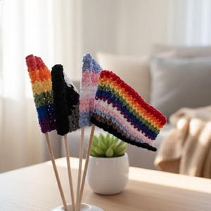 May include: A collection of five small, handmade crochet flags on wooden dowels. The flags display various rainbow and color block patterns, including the Pride flag and the Transgender Pride flag. The flags are arranged in a white ceramic holder on a light-colored table.