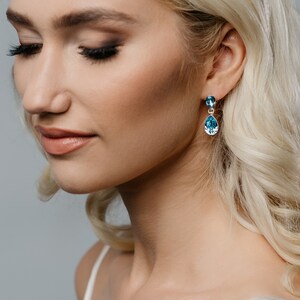 Puede incluir: Elegantes pendientes colgantes con cristales azules en forma de lágrima. Los pendientes están engarzados en un metal plateado y cuelgan con gracia. El cristal superior es redondo, mientras que el inferior es una piedra más grande en forma de pera. Una joya clásica.