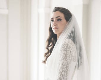 Op de afbeelding: Een bruid in een witte kanten jurk met een lange sluier, die een boeket witte bloemen vasthoudt. Ze staat op een trap in een witte kamer.