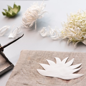 May include: A still-life image of crafting supplies. White paper flower petals and a finished flower are arranged with a soldering iron and a linen cloth. The scene suggests a handmade floral craft project.