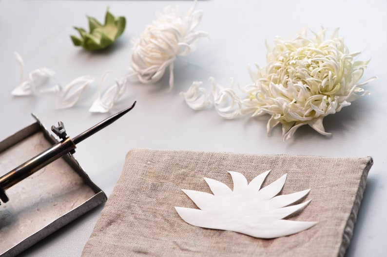 May include: A still-life image of crafting supplies. White paper flower petals and a finished flower are arranged with a soldering iron and a linen cloth. The scene suggests a handmade floral craft project.