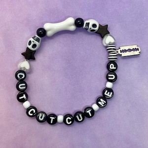 Peut inclure: Bracelet en perles noires et blanches avec les mots "CUT ME UP" et des breloques en forme de crâne et d'os.