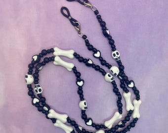 May include: Black and white beaded glasses chain with skull and bone charms. The chain has black heart charms and silver metal clasps.