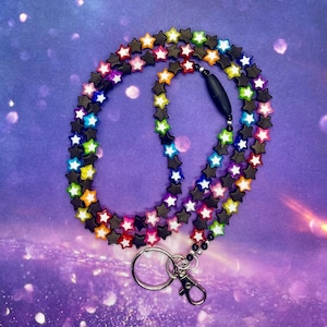 Op de afbeelding: Een sleutelkoord met zwarte stervormige kralen, elk met een kleurrijke ster in het midden. Het sleutelkoord heeft een zilveren sluiting en een sleutelring. De achtergrond is een paarse en blauwe gradiënt met glitter.