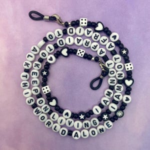 May include: Black and white beaded glasses chain with the words "I am not afraid to keep on living alone" and dice and heart charms.