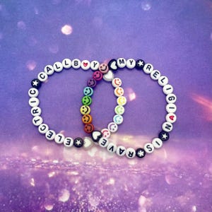 Könnte beinhalten: Zwei weiße Perlenarmbänder mit schwarzer Beschriftung. Ein Armband lautet "Call My Religion" und das andere "Electric Rave". Beide Armbänder haben Smiley-Perlen und Herzperlen.