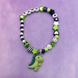 Può includere: Un braccialetto di perline colorato con un ciondolo a forma di dinosauro. Il braccialetto presenta perline bianche con le lettere "RAWR", perline verdi a forma di stella e un ciondolo a forma di dinosauro verde. Altre perline includono strisce bianche e nere, teschi e faccine.
