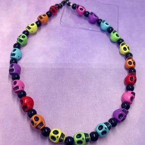 Op de afbeelding: Een kleurrijke telefoonbedel met een herhalend patroon van kleine schedel kralen in een regenboog van kleuren, waaronder rood, oranje, geel, groen, blauw, paars en roze. De schedels worden afgewisseld met zwarte kralen.