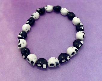 May include: A bracelet made of black and white skull beads. The beads are skull-shaped, with alternating black and white colours. The bracelet is circular in shape. The background is a solid purple colour.