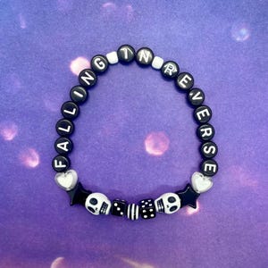 May include: A black beaded bracelet with white lettered beads spelling "FALLING INVERSE". The bracelet includes skull and star charms, and heart-shaped beads. The background is a purple gradient.