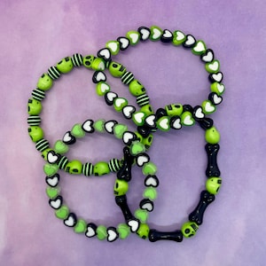 Può includere: Tre braccialetti con perline verdi e nere. Un braccialetto ha cuori bianchi, uno ha perline a strisce bianche e nere e l'ultimo ha perline nere a forma di osso con teschi.