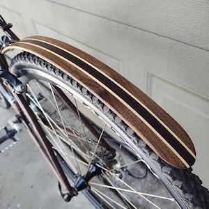 May include: A close-up of a wooden bicycle fender with a brown and black striped design. The fender is attached to a bicycle wheel with a black tire.