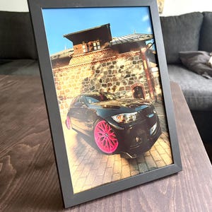 Puede incluir: Un marco de fotos negro que contiene una fotografía de un sedán negro con llantas de color rosa brillante de múltiples radios estacionado sobre una superficie de ladrillo frente a un edificio de piedra con techo marrón.