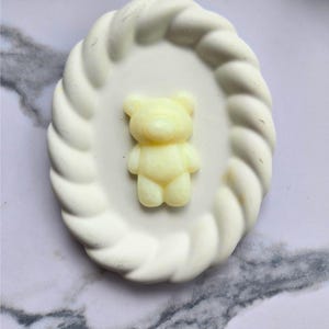 May include: A pale yellow bear-shaped object centred on a white oval tray. The tray features a decorative rope-style border around its edge. The bear figure has a smooth, rounded appearance, while the tray has a textured, twisted rim design.