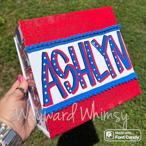 May include: A rectangular box with a red glittery finish featuring a white center panel. The name ASHLYN is written in large blue letters with red outlines and white dots. The panel is bordered by blue pom-pom trim on the top and bottom edges.