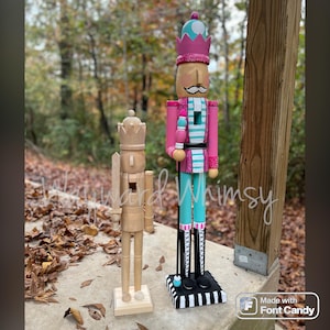 May include: Two wooden nutcrackers. One is natural wood with a crown and sword. The other is painted pink, teal, and white, with a mustache and a tall crown. The larger nutcracker stands on a black and white striped base.