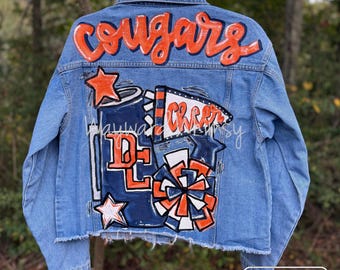 May include: Light blue denim jacket with hand-painted orange "Cougars" lettering across the shoulders. The back features a cheerleading design with stars, pom-poms, and the word "Cheer".