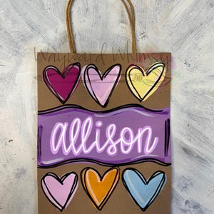 May include: A brown paper gift bag with a jute handle. The bag features a purple banner with the name "allison" in white script, and six colorful hearts in shades of pink, yellow, orange, and blue.