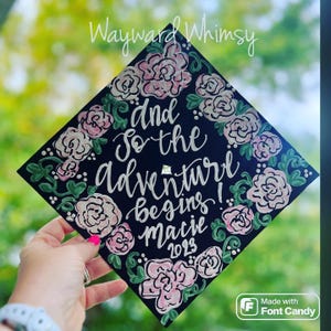 May include: Black graduation cap decorated with pink and green flowers and the text "And so the adventure begins! Macie 2023".