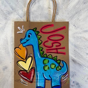 May include: A brown paper gift bag with a blue dinosaur illustration and the name "JOSH" in pink. The dinosaur has green spikes and spots, with three colorful hearts. The bag has a brown twisted paper handle.