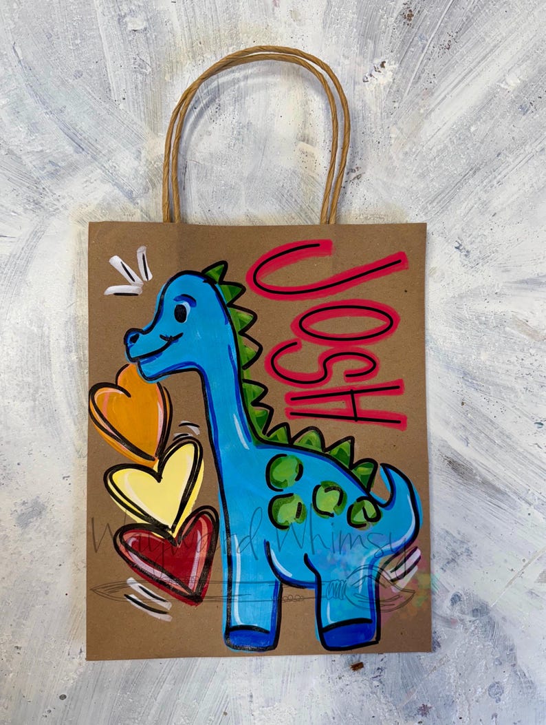 May include: A brown paper gift bag with a blue dinosaur illustration and the name "JOSH" in pink. The dinosaur has green spikes and spots, with three colorful hearts. The bag has a brown twisted paper handle.