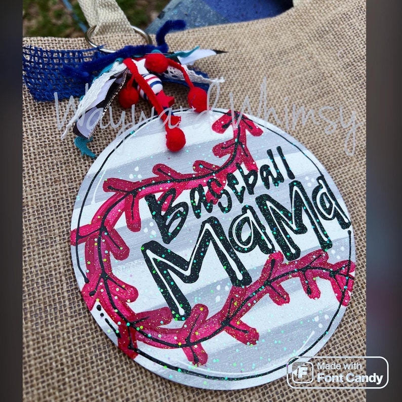 Może przedstawiać: Okrągły znak w biało-szare paski z czerwonym wzorem szw&oacute;w baseballowych i napisem "Baseball Mama" czarnym pismem. Znak ma błyszczące wykończenie.