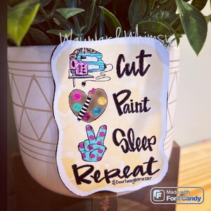 May include: A yellow background with black text that reads "Cut, Paint, Sleep, Repeat" with a hashtag that reads "#doorhangammaker". The image also includes a small illustration of a sewing machine, a paint palette, and a peace sign.
