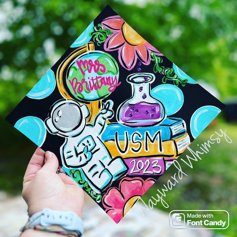 May include: A black graduation cap painted with a colorful design. The design includes a globe, an astronaut, a beaker, books, flowers, and the text "Mrs. Brittany USM 2023".