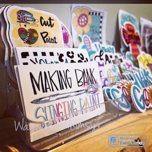 May include: A clear plastic display stand filled with colorful stickers and cards. The cards have various designs, including a paintbrush with the text "Making Bank" and "Slinging Paint".