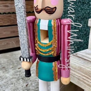 May include: Wooden nutcracker figurine with a crown, mustache, and a silver glitter sword. The nutcracker is painted in shades of pink, teal, and gold, with black boots and a teal base. A decorative item for the holidays.
