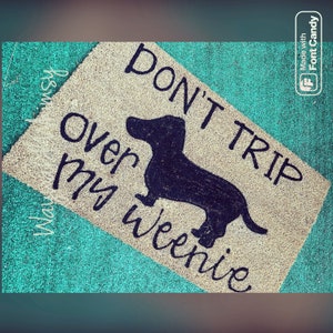 May include: A rectangular doormat with the text "DON'T TRIP Over my Weenie" and a black silhouette of a dachshund. The mat is beige with a teal border.