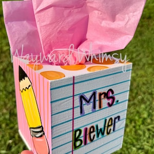 May include: A square wooden tissue box cover painted to resemble a piece of lined notebook paper with a yellow pencil illustration on the side. The front features the text Mrs. Brewer in colorful lettering. Pink tissue paper extends from the top.