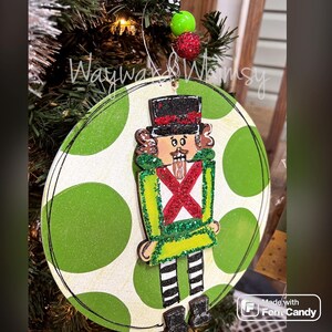 May include: A round wooden ornament with a green and white polka dot pattern. A painted nutcracker figure is attached to the ornament. The nutcracker is wearing a green and red suit with a black hat and white boots. The ornament has a string for hanging.