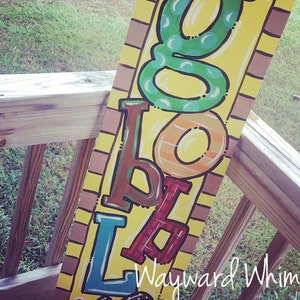 May include: A wooden sign with the word "GO" painted in green, "TO" painted in orange, "SCHOOL" painted in red, blue, and brown, and a turkey painted at the bottom. The sign has a brown brick pattern on the side.