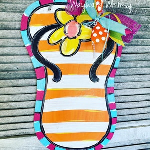 May include: A wooden flip-flop shaped door hanger with orange and white stripes. It features a yellow flower with a pink center, black straps, and a decorative bow. The edges are outlined in pink, blue, and black. The text "Wayward Whimsy" is visible.