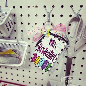 May include: A white tag with black stripes and the text "Mrs. Bridental" in pink and purple lettering. The tag is decorated with colorful, rainbow-colored crayons and is attached to a key ring with a pink and white ribbon.
