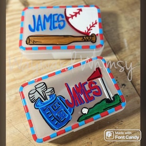 May include: Two clear plastic boxes with red, white, and blue trim. One box features a baseball and bat design with the name "JAMES" in blue. The other box has a golf-themed design with the name "JAMES" in red.