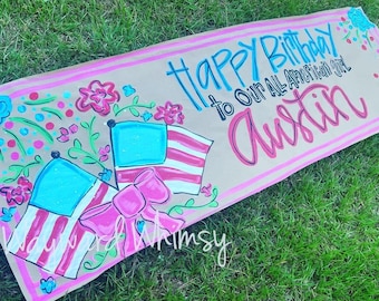 Könnte beinhalten: Ein braunes Papierbanner mit rosa und blauen Streifen an den Rändern. Das Banner hat eine handgemalte Botschaft, die "Happy Birthday to our All American Girl Austin" lautet. Das Banner ist mit handgemalten Blumen, Sternen und Flaggen verziert.