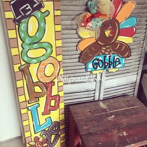 May include: A wooden sign with the word "Gobble" spelled out in colorful letters. The sign is decorated with a turkey and a pilgrim hat. The sign is on a wooden background.