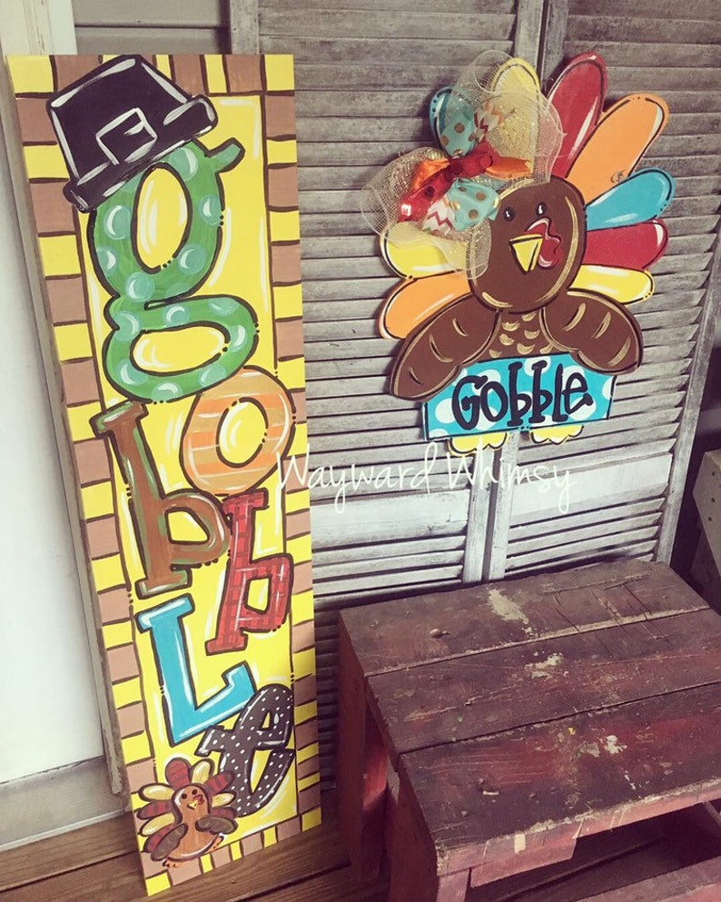 May include: A wooden sign with the word "Gobble" spelled out in colorful letters. The sign is decorated with a turkey and a pilgrim hat. The sign is on a wooden background.